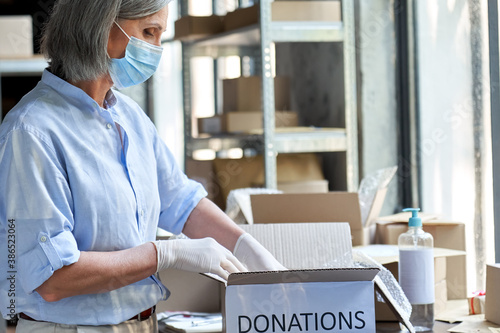 Mature female warehouse worker volunteer wearing face mask working in shipping delivery charitable stock organization packing donations box. Covid 19 coronavirus donating and volunteering concept.