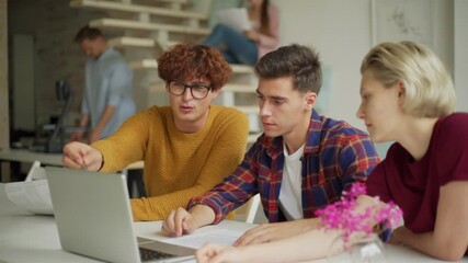 Medium shot of group of three young business people sitting at table, looking at laptop screen and discussing project