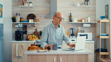 Daily life of senior man in kitchen during breakfast using laptop holding a cup of coffee. Elderly retired person working from home, telecommuting using remote internet job online communication on