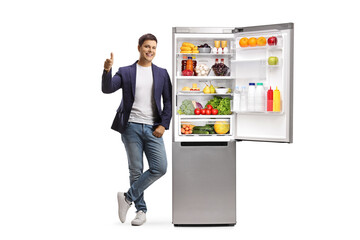 Sticker - Full length portrait of a young man leaning on a full fridge and showing thumbs up