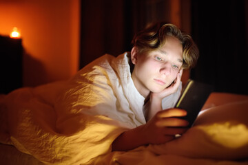 Teenager boy chatting by phone, writing in blog or reading social networks instead sleeping. Young guy laying in bed and watching clips by smartphone. Overuse and addiction kids from gadgets.