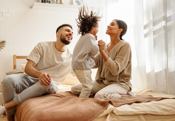 happy diverse family mom, dad and child laughing, playing and jumping in bed at home.