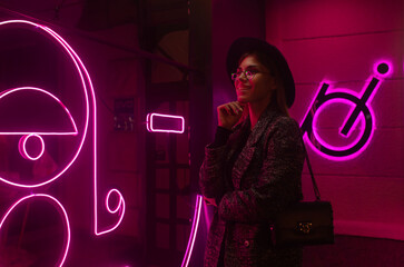 Stylish woman in trendy coat and eyewear looking up enjoying nightlife in city. Gorgeous fashion hipster girl in jacket standing outdoors on street with neon city illumination.