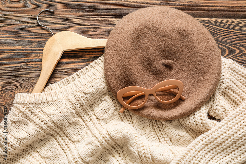 Clothes hanger with sweater, glasses and cap on wooden background