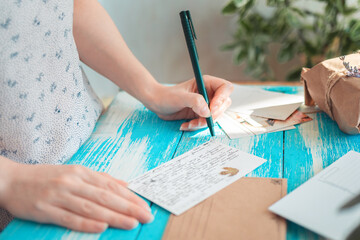 A woman writes a letter on a postcard. Hands holding a pen close-up. Blue wooden table with a parcel in the background. The concept of postcrossing