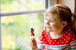 © Irina Schmidt - Cute little toddler girl sitting by window on rainy day during pandemic coronavirus quarantine. Adorable child eating and licking rainbow colored lollipop
