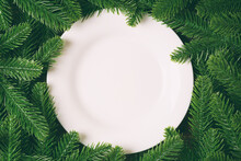 Top View Of White Plate Surrounded With Fir Tree Branches. Christmas Dinner Concept With Copy Space