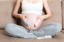 Beautiful Pregnant Young Woman Holding A Pair Of Cute Baby Socks On Her Tummy, Lying On Sofa At Home