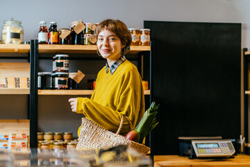 Wall Mural - Minimalist vegan style girl with wicker bag and reusable glass coffee cup on background of interior of zero waste shop. Woman doing shopping without plastic packaging in plastic free grocery store.