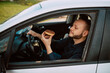 © Dragica - Side view of a young caucasian businessman driving a car on the way to work and eating a delicious doughnut with chocolate cream. Sweet breakfast in the car
