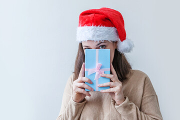 Wall Mural - Beautiful girl in red Santa Claus hat holding blue gift box in hand isolated on white background. Young woman portrait, true emotions. Happy Christmas and New Year holidays concept.