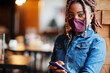 © AS Photo Family - Stylish african american woman with dreadlocks afro hair, wear jeans jacket and face protect  mask at restaurant, hold cellphone. New normal life after coronavirus epidemic.