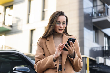 Wall Mural - Business woman in autumn coat browsing internet on smartphone outdoors. Smiling lady with long brown hair using modern gadgets on street.