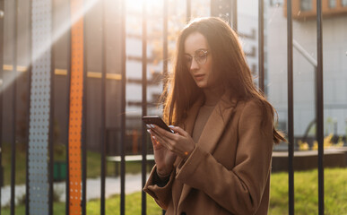 Wall Mural - Smiling curly businesswoman wearing trendy sunglasses walks down the central city street and uses her phone. Businesswoman holding cell phone.