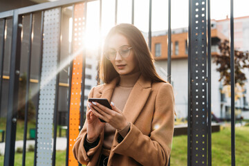 Wall Mural - Business woman curly woman wearing trendy sunglasses walks down the central city street and uses her phone. Pretty summer woman in brown jacket walks down the street looking at her mobile phone.