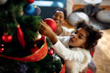 Small black girl decorating Christmas tree at home.