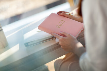 Female hands holding pink coral coloured leather diary 2021 while sitting near a window at cafe