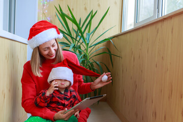 christmas online congratulations: a woman with a girl in red Santa hats congratulating family or friends with a tablet. the mother and daughter celebrates the new year in isolation. Winter holidays