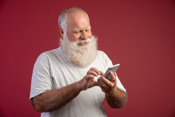 Smiling elderly bearded man using smartphone on red background. Senior man using wireless wi-fi internet connection on smart phone. Happy old man looking at screen on smartphone.