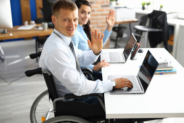 Businessman in wheelchair and businesswoman at table are smiling and raised their hands in greeting. Positive work mood in people with disabilities concept