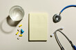 © uaPieceofCake - Top view of blue stethoscope, glass of water, yellow and blue capsule pills, blank sheet of noteebok.Empty space for prescription.Hard light