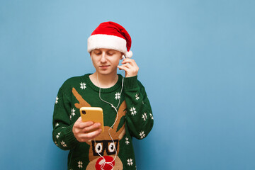 Wall Mural - Handsome young man in sweater with deer and santa hat stands on blue background with smartphone in hand and listens to music in headphones. Guy in a sweater and a santa hat listens to Christmas music