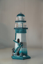 A Figurine In The Form Of A Lighthouse With A Large Anchor In The Foreground. Blurred Background.
