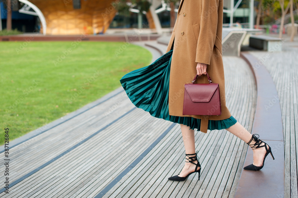 Fashionable young woman wearing green pleated midi skirt, sweater, high  heel shoes, beige wool coat and holding burgundy handbag in hand on the  city street. Street style. Stock Photo | Adobe Stock