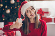 Pretty woman raise hand resent thumb up toothy smile wear santa headwear red sweater in decorated x-mas living room indoors