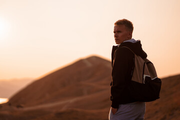 Canvas Print - Picturesque mountain landscape at sunset with the sea on the horizon, rear view of young blond man with backpack stands on the slope in the orange rays of the sunset