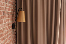 Stylish Small Golden Lamp Sconce Installed On Red Brick Wall Against Brown Window Curtain In Contemporary Apartment Room Closeup. Interior Design Details.