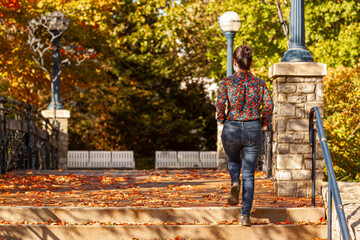 Wall Mural - A young caucasian woman wearing floral shirt and jeans is climbing stairs in a city park. It is a beautiful autumn scene with colorful leaves fallen on ground. A concept image for fall, loneliness,