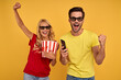© Roman - Joyful young couple friends guy girl in 3d glasses isolated on yellow background. People in cinema concept. Watching movie film, hold bucket of popcorn, mobile phone doing winner gesture