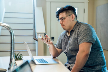 Wall Mural - man working from home at kitchen counter with laptop computer looking at mobile phone