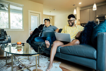 Wall Mural - happy gay couple working from home with laptops on couch with their australian shepherd