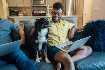 Wall Mural - happy gay couple working from home with laptops on couch with their australian shepherd