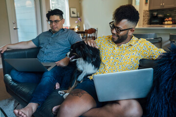 Wall Mural - happy gay couple working from home with laptop computers on couch with their pet dog