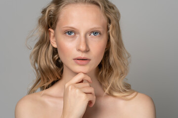 Close up of beautiful young woman face with blue eyes, curly natural blonde hair, has no makeup, touching her chin, standing shirtless with bare shoulders, looking at camera. Studio grey background.