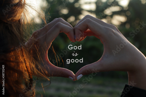 Young woman hand making love sign with message - God is good. Hope and believe in God concept. Fingers with heart shape gesture against sunset light on green background in field.