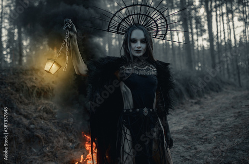 Woman in image of witch stands with glowing lamp against black smoke and fire in forest.