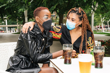 Two multiracial friends on a bar terrace having a drink and hugging each other with face masks