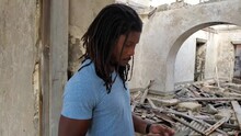 Black African Caribbean Dreadlock Rasta Man Looks At Damage And Call Texts On Phone For Help In Home Natural Disaster Scene - Hurricane, Storm, Tornado Cyclone