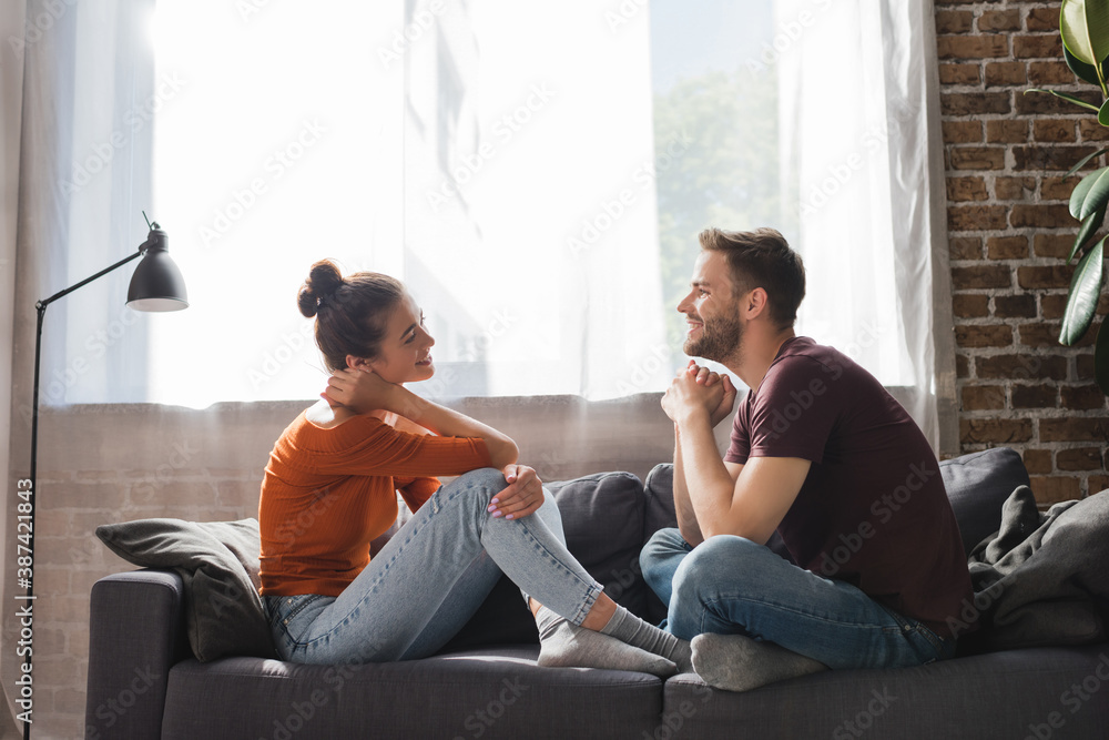 side view of young couple sitting on sofa and talking while looking at each other - obrazy, fototapety, plakaty 