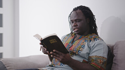 African Man in traditional dress with rosary reading the Holy Bible sitting on the sofa. High quality photo