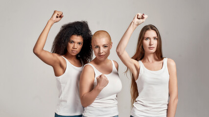 Portrait of three young diverse women wearing white shirts having confident look while showing, raising clenched fist, posing together isolated over grey background