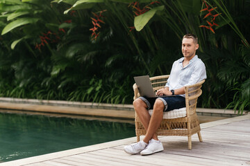 Young man using a laptop computer in a garden with a swimming pool. business, study, freelance