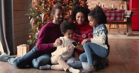 Wall Mural - Full african family sit on warm floor in cozy modern living room spend winter holidays on internet using smartphone enjoy new trendy app taking selfie having fun together near decorated Christmas tree