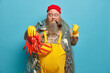 © WHstudio Leushin N - Serious bearded boat captain clenches fist as warns about revenge holds sea creature keeps smoking pipe in mouh carries fishing net stands against blue background. Sea cruise or travel concept