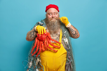 Positive bearded boatswain curls mustache and looks away happily wears rubber gloves and overalls has tattoed arms holds red octopus isolated over blue background. Sea travel and fishing concept