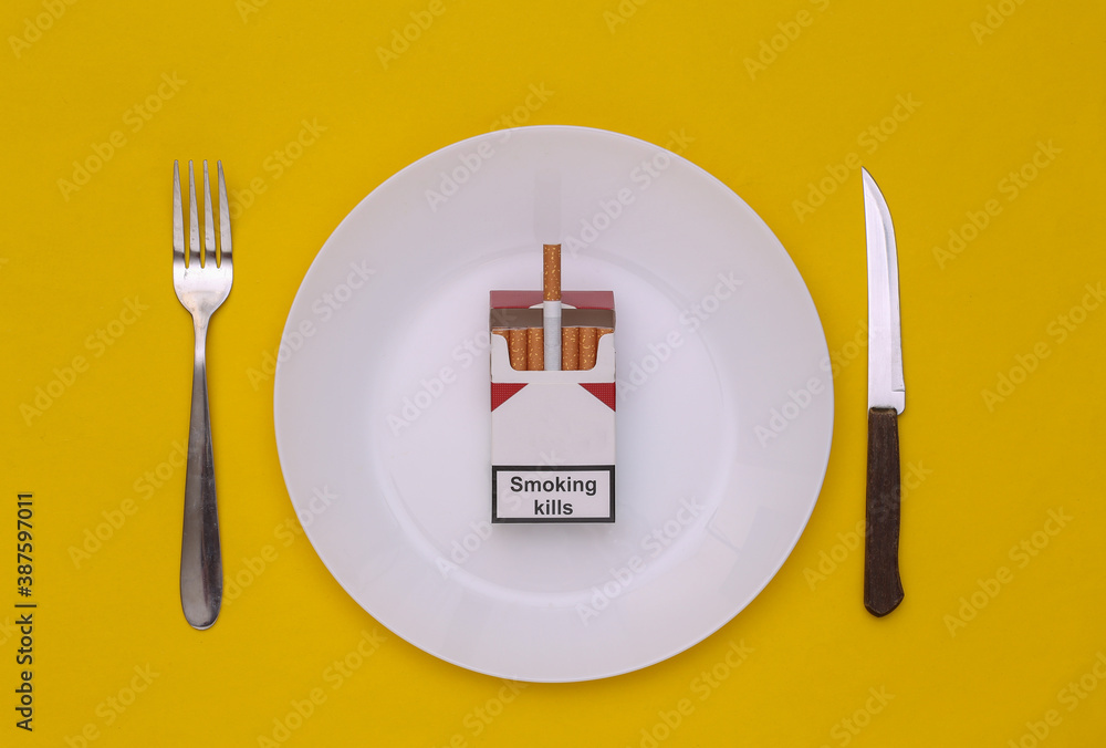 plate of cigarettes Pack of cigarettes in plate, fork and knife on yellow background. Food, bad  habits concept. Top view. Stock Photo